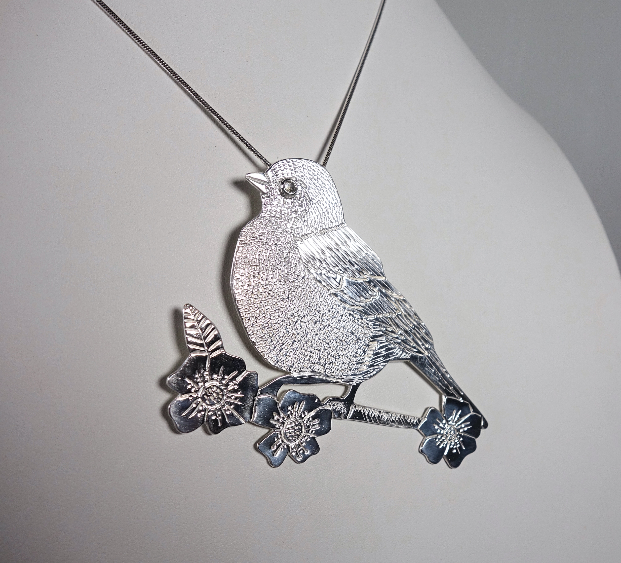 Pendentif oiseau Madame M en argent massif et péristérite, vue de profil sur buste blanc, à la lumière artificielle