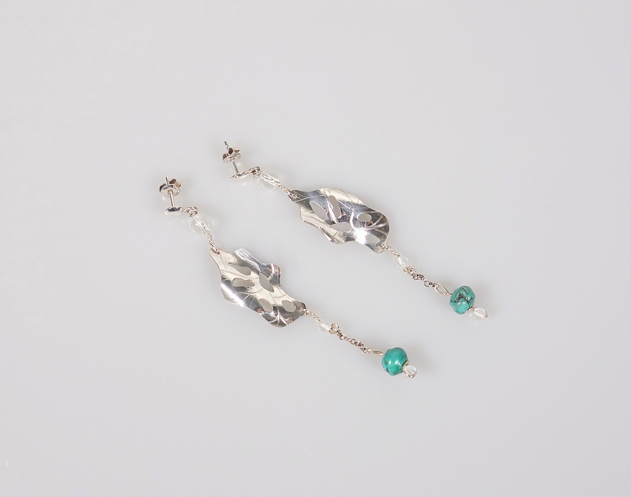 Boucles doreilles pendantes &quot;Cheyennes&quot; en argent massif et perles en turquoise et cristal de roche, vue de biais sur fond blanc à la lumière électrique
