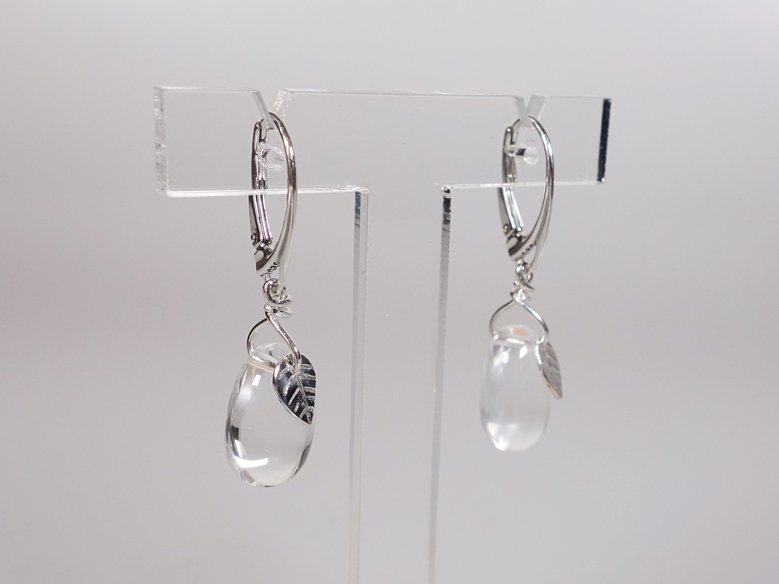 Boucles doreilles gouttes en cristal de roche et feuille, vue de biais, sur présentoir et fond blanc à la lumière artificielle
