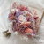 Un bouquet de mariée bohème composé de roses délicates en nuances de rose, entourées de diverses fleurs et feuillages dans des tons de rose, violet et blanc