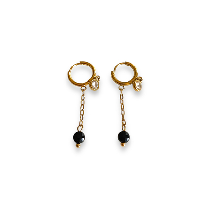 boucles d oreilles petites creoles dorees avec pampille sertie d un strass et chaine avec pierre naturelle de couleur noire a l extremite exposees fond blanc tara