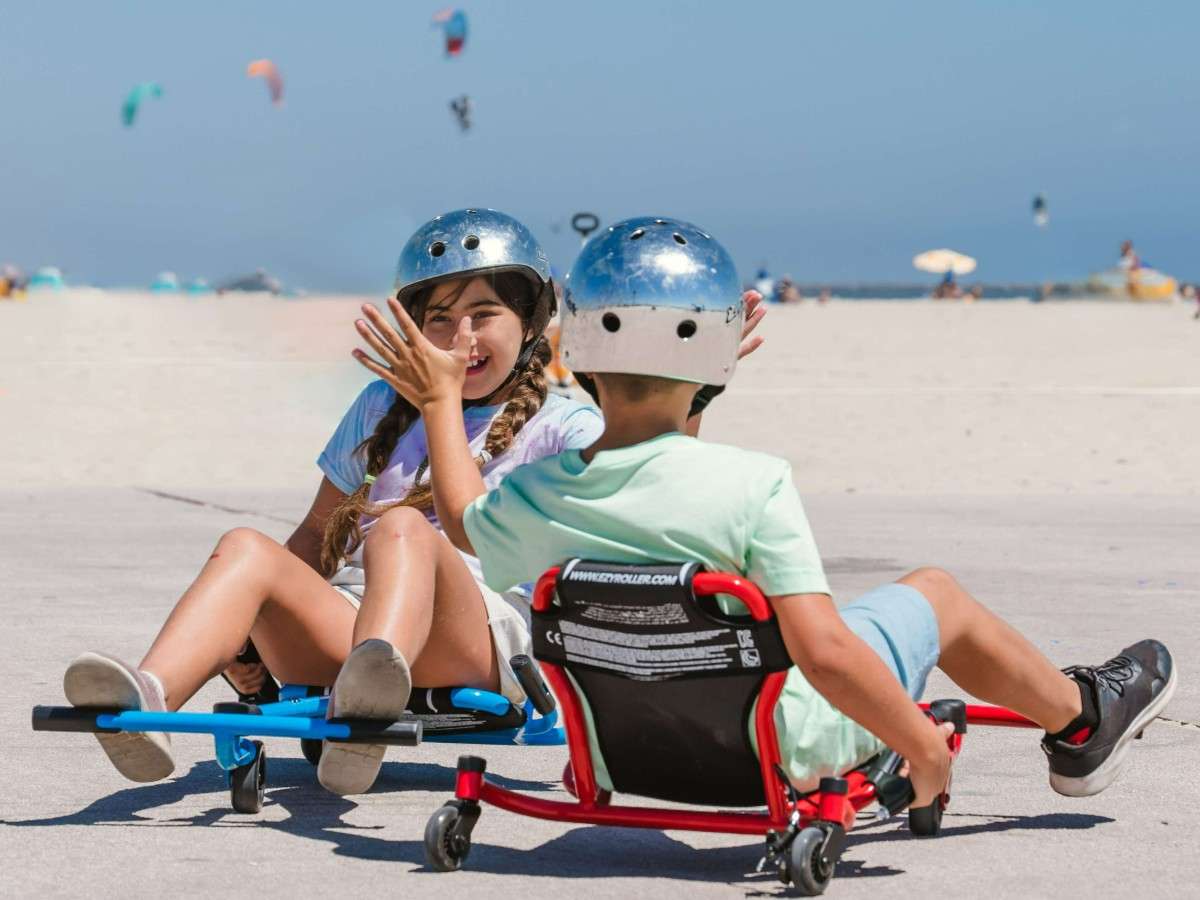 Deux enfants casqués jouent ensemble sur des Ezyroller Spinner X, l’un en rouge et l’autre en bleu, sur une zone dégagée en bord de plage.
