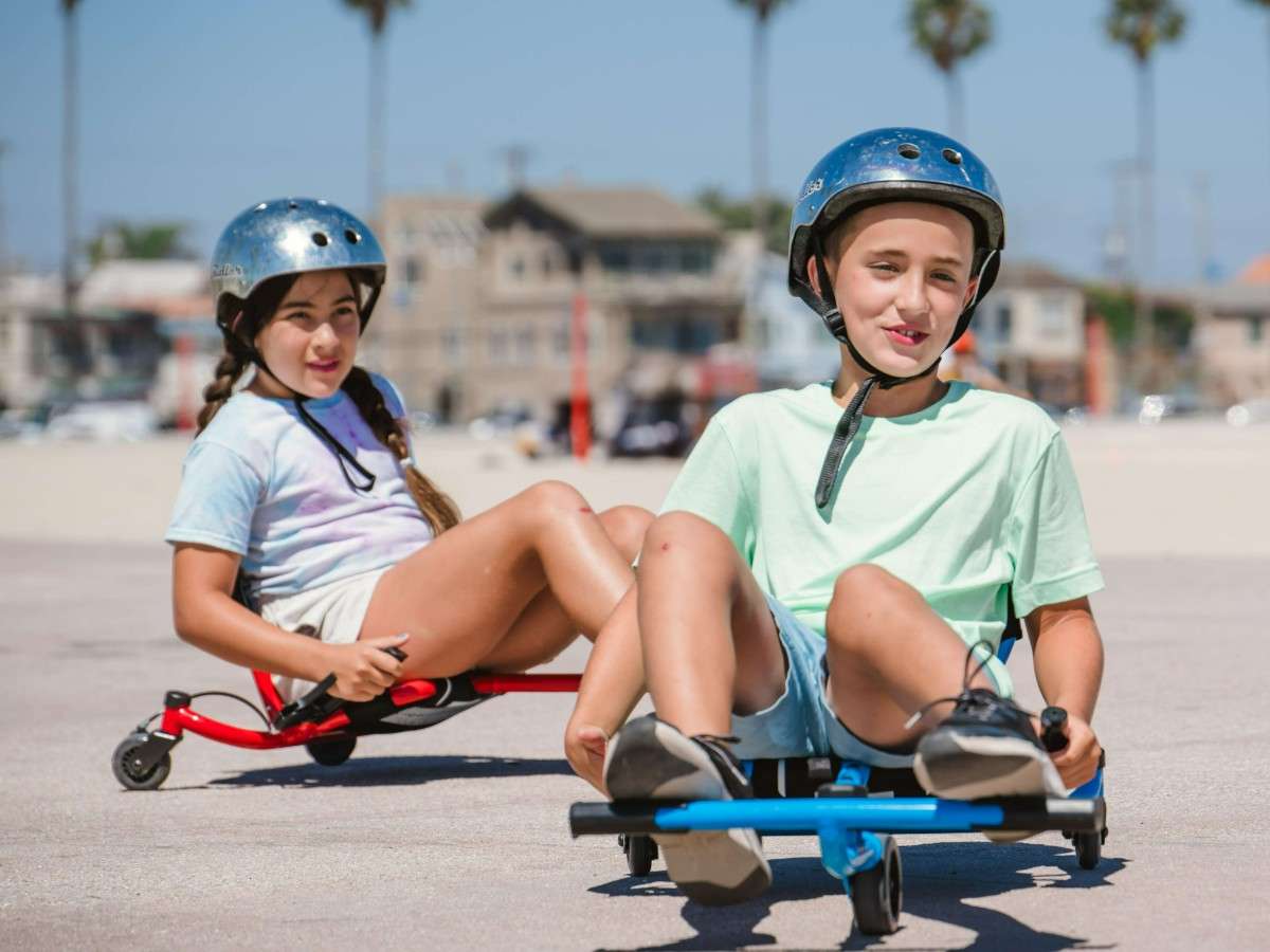 Deux enfants casqués roulent sur des Ezyroller Spinner X, l’un en bleu et l’autre en rouge, en jouant sur une zone dégagée en plein air.