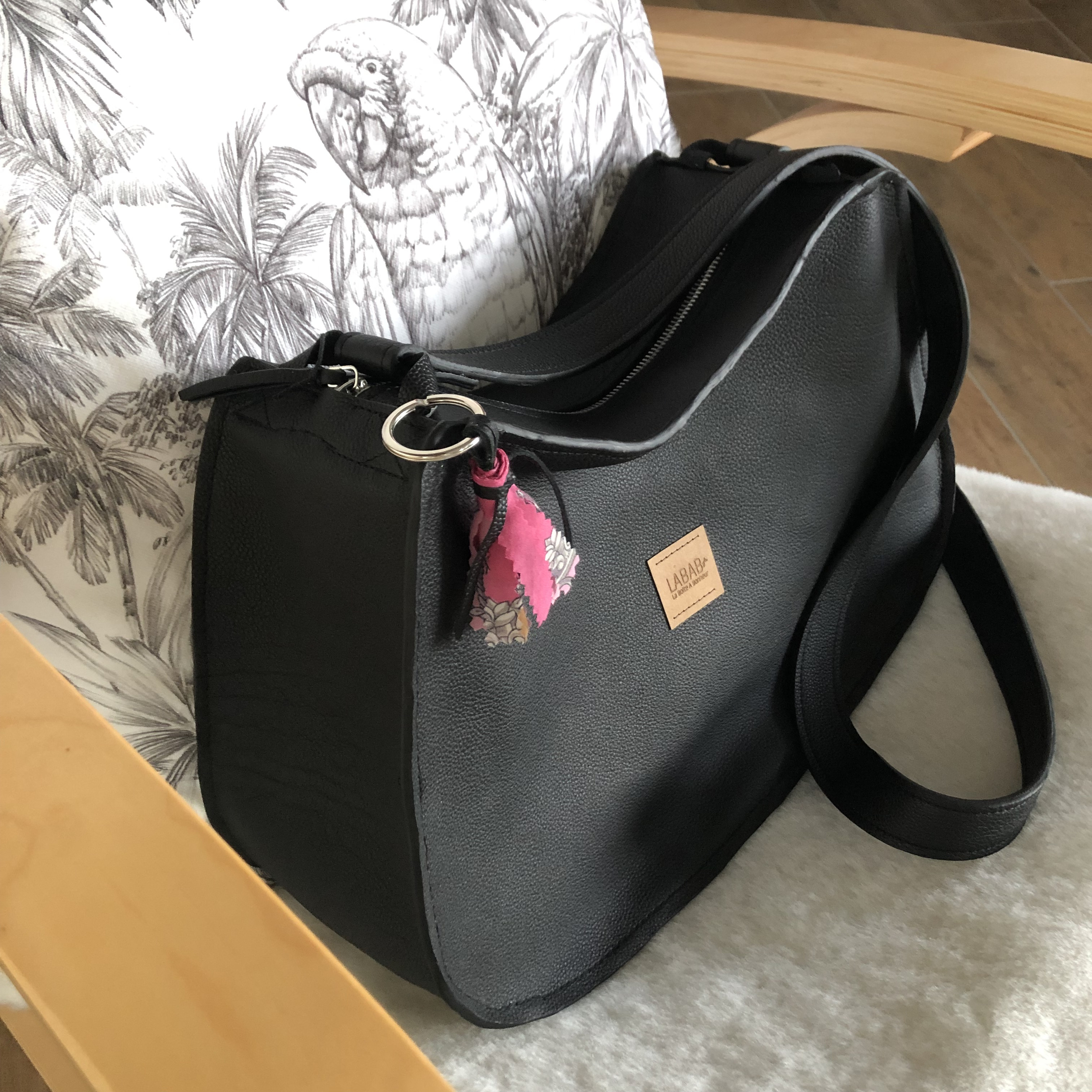 sac à main cuir noir et bijoux de sac pompon en foulard rose posé sur un fauteuil montrant volumes du sac