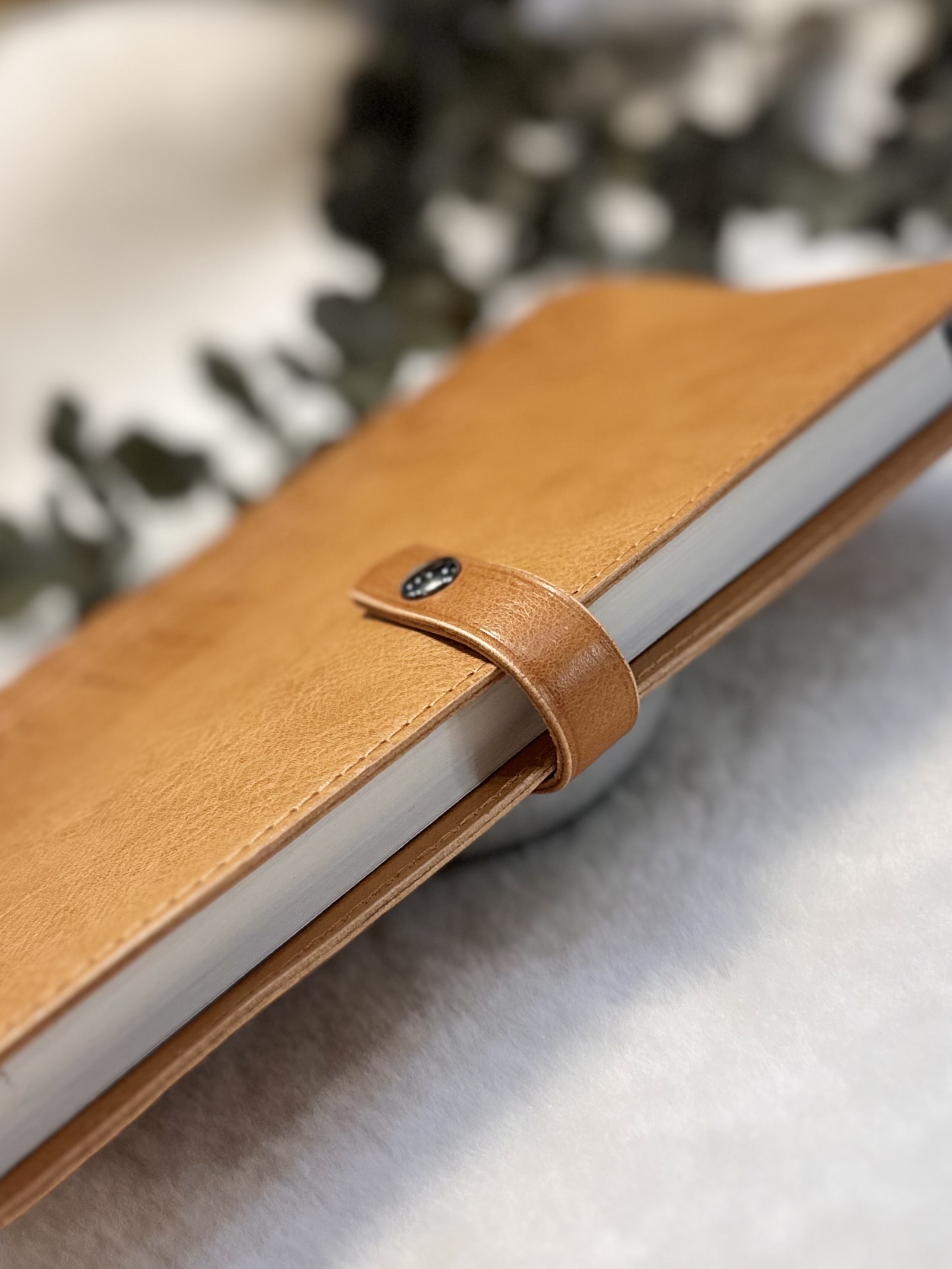 Protège-carnet en cuir camel avec cahier dedans vu de profil mettant en évidence les tranches et la patte en cuir qui ferme le protège-carnet à laide dun bouton pression en laiton