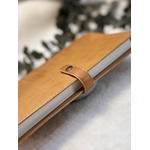 Protège-carnet en cuir camel avec cahier dedans vu de profil mettant en évidence les tranches et la patte en cuir qui ferme le protège-carnet à laide dun bouton pression en laiton