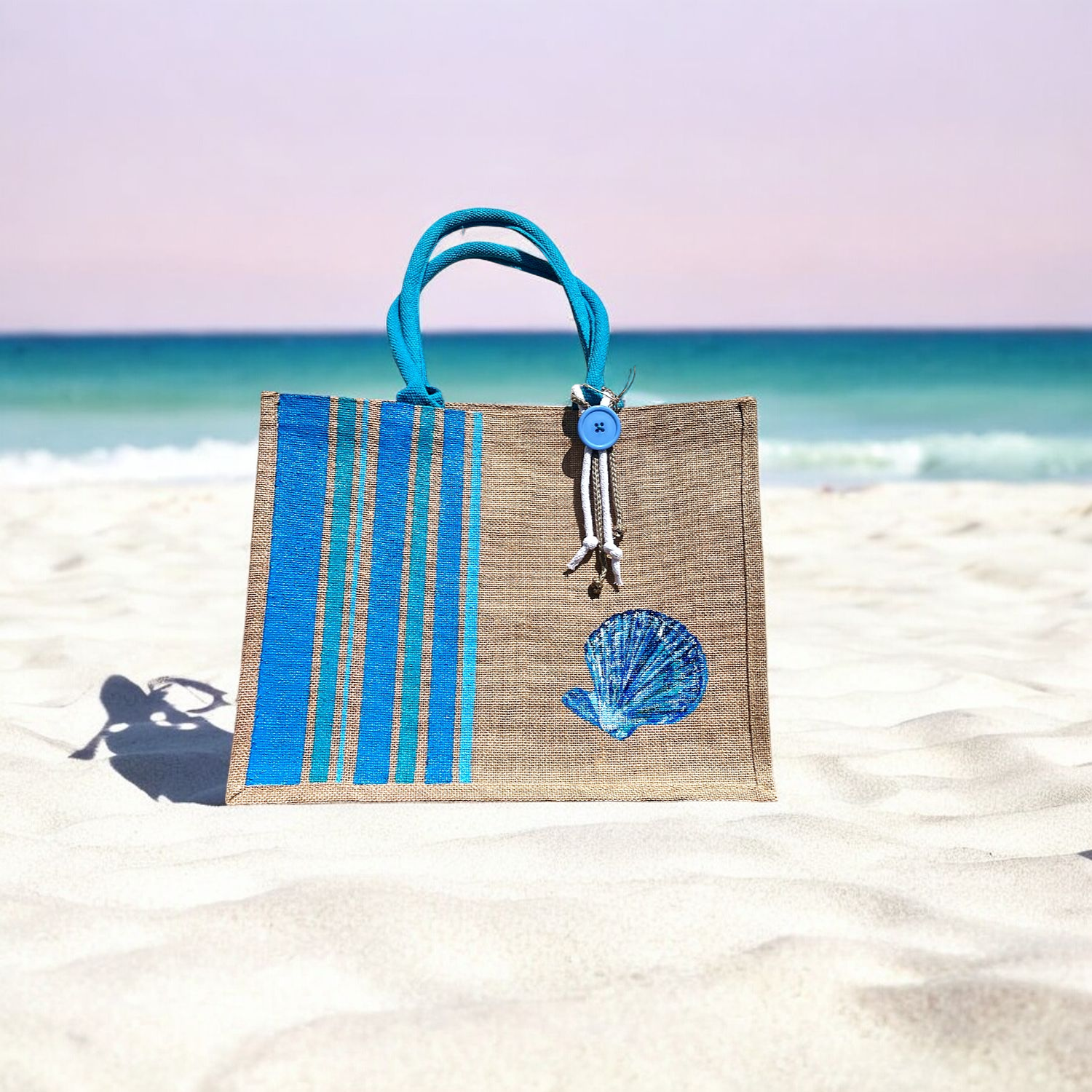 grand sac de plage en jute avec des bandes bayadère bleus et un coquillage peints à la main par Elle et No. Un colifichet original est attaché à la anse.