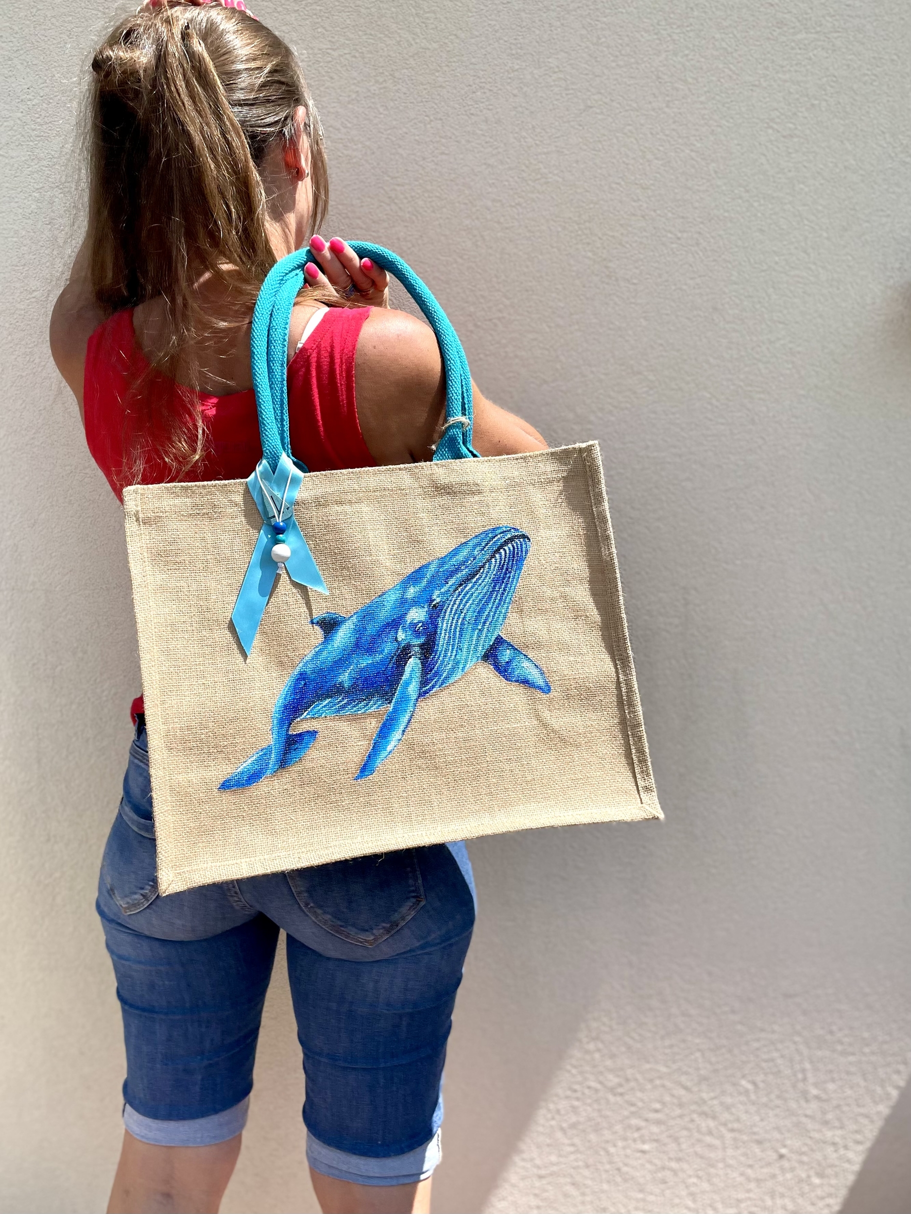 sac de plage en jute avec une baleine bleue peinte à la main par Elles et No., présenté porté par une femme de dos