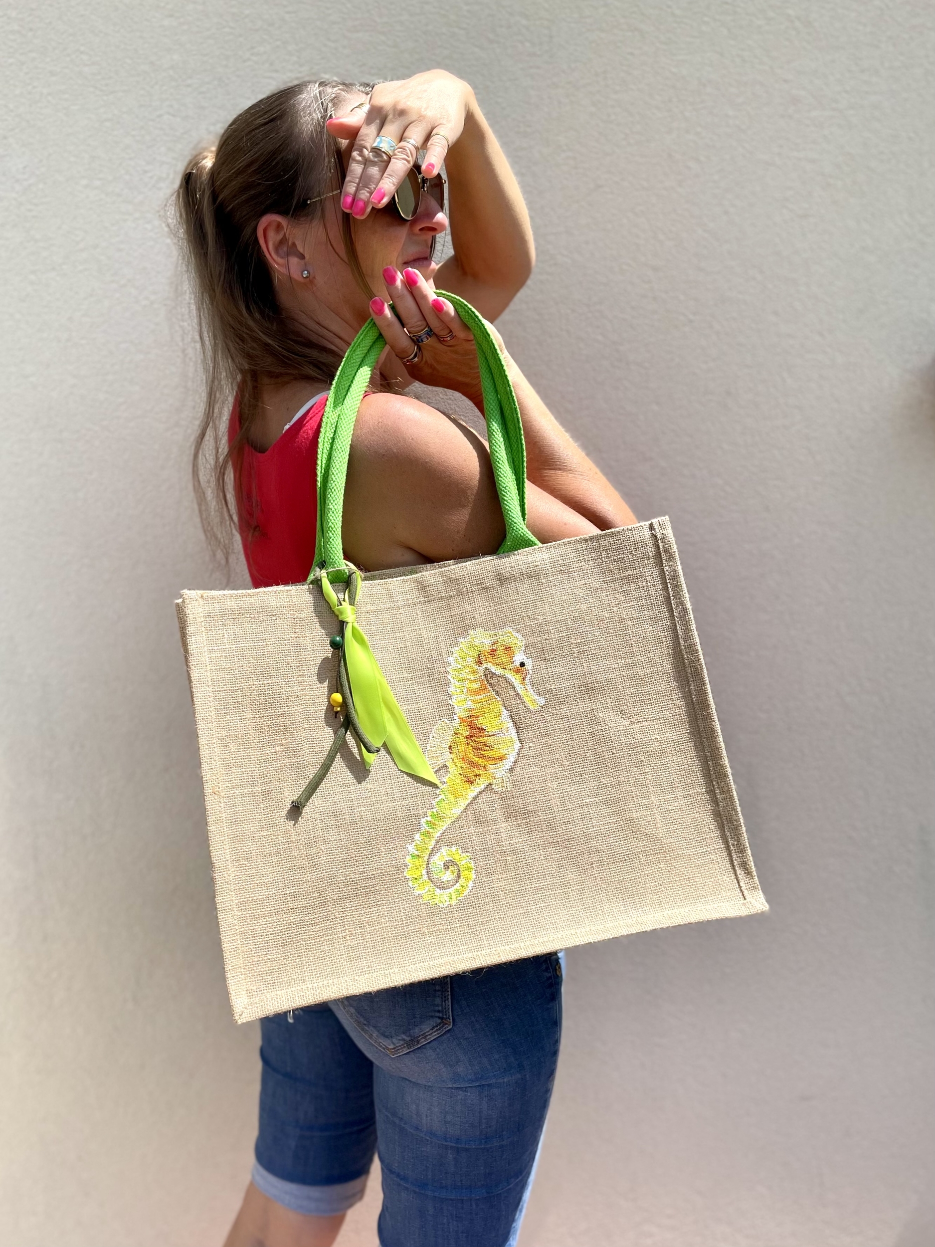 sac de plage en jute à anses vertes avec un hippocampe dans les tons jaune et vert peint à la main par Elles et No., présenté porté par une femme