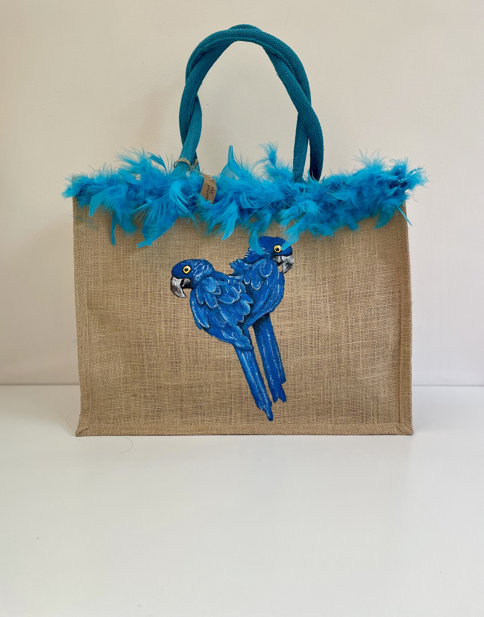 Sac de plage en jute avec des perroquets bleus peints à la main par Elles et No. et un boa en plumes collés sur le bord du sac