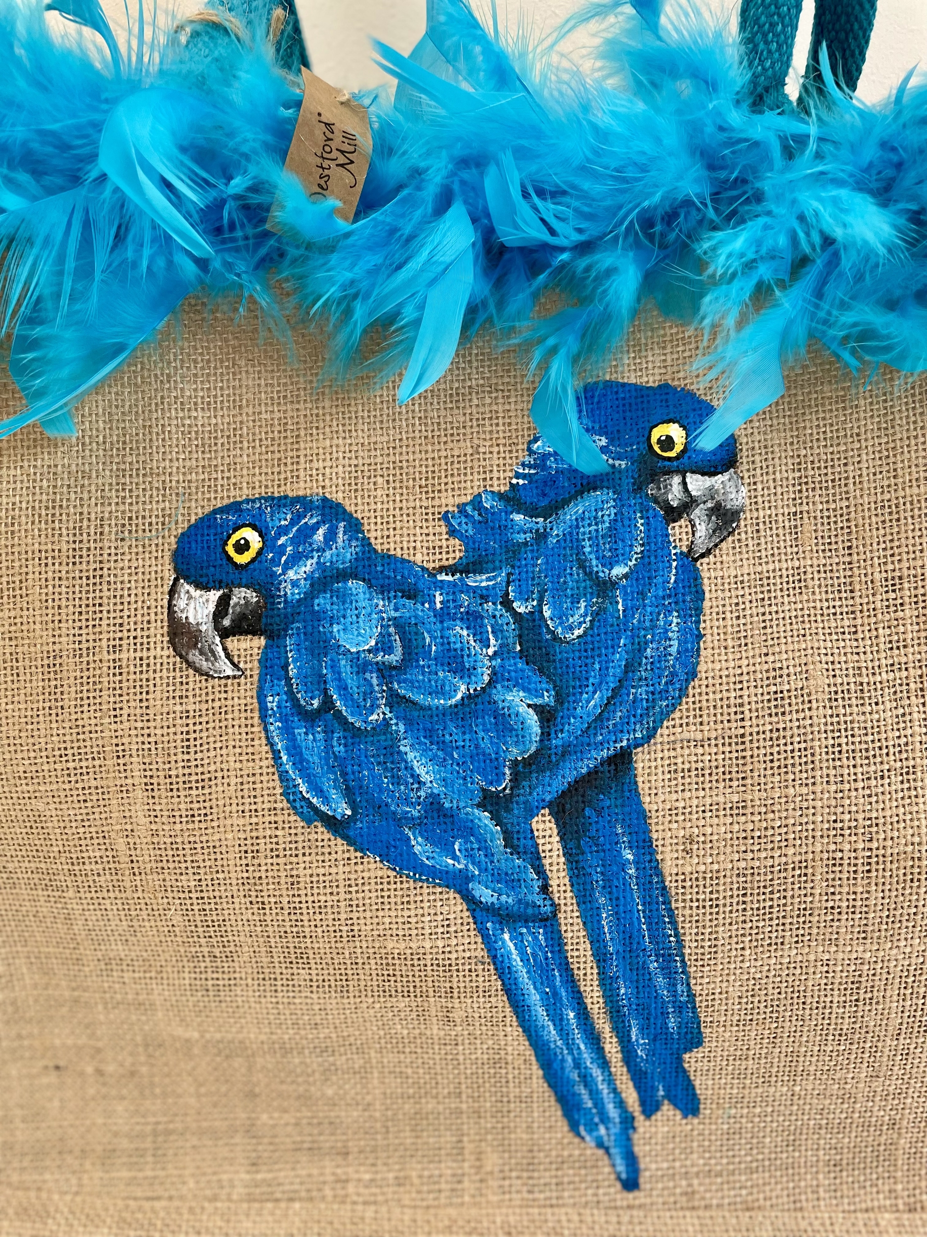 Sac de plage en jute avec des perroquets bleus peints à la main par Elles et No. et un boa en plumes collés sur le bord du sac