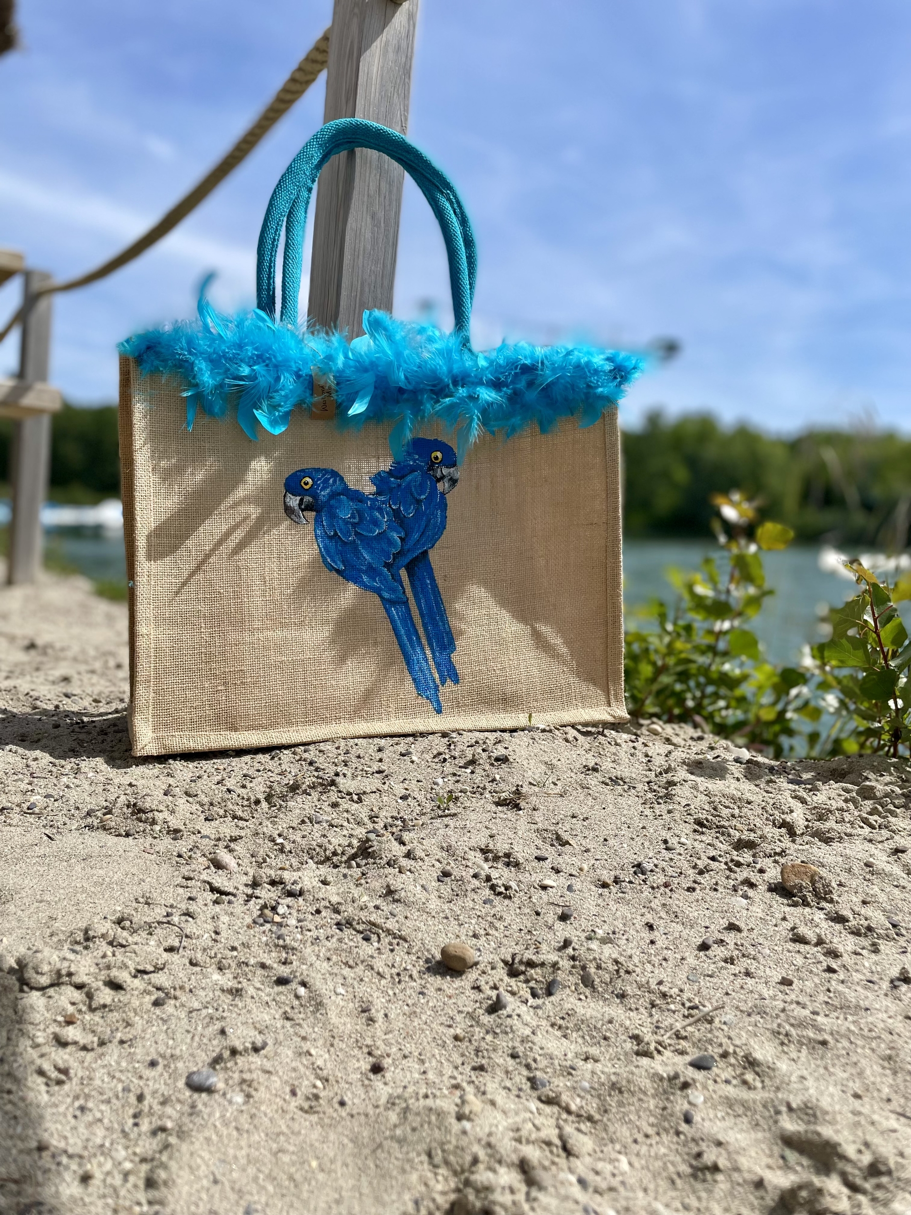 Sac de plage en jute avec des perroquets bleus peints à la main par Elles et No. et un boa en plumes collés sur le bord du sac