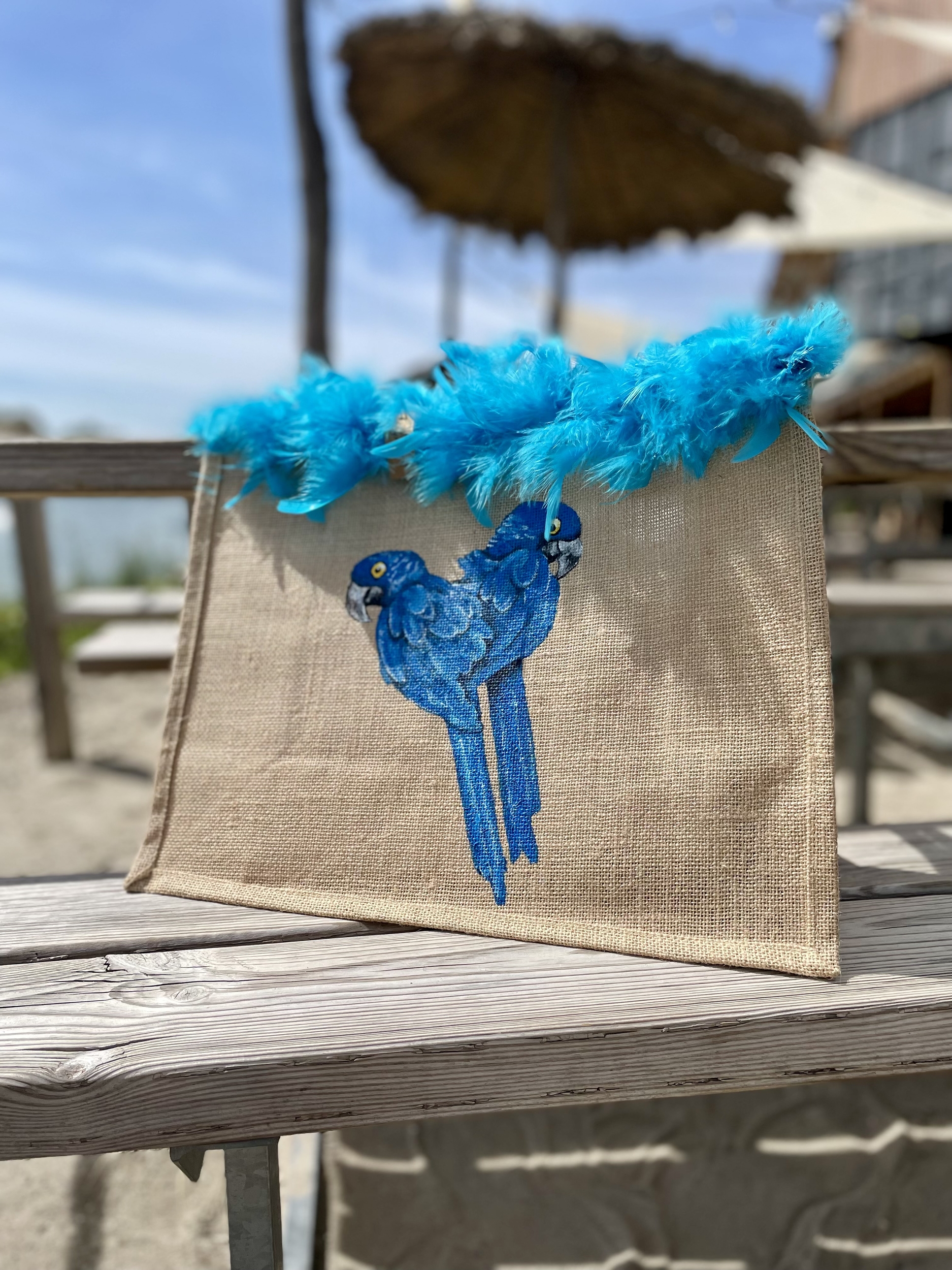 Sac de plage en jute avec des perroquets bleus peints à la main par Elles et No. et un boa en plumes collés sur le bord du sac