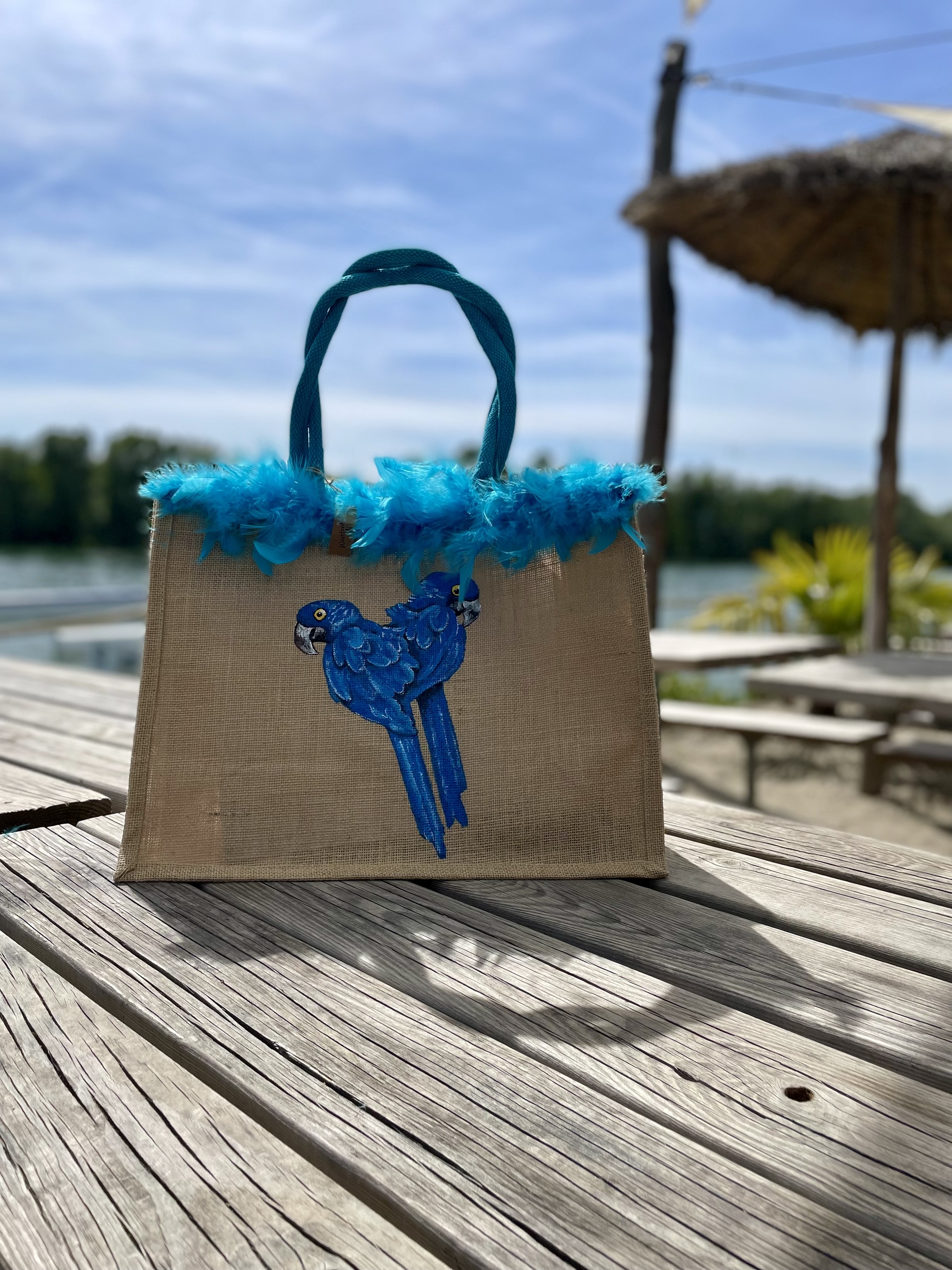 Sac de plage en jute avec des perroquets bleus peints à la main par Elles et No. et un boa en plumes collés sur le bord du sac