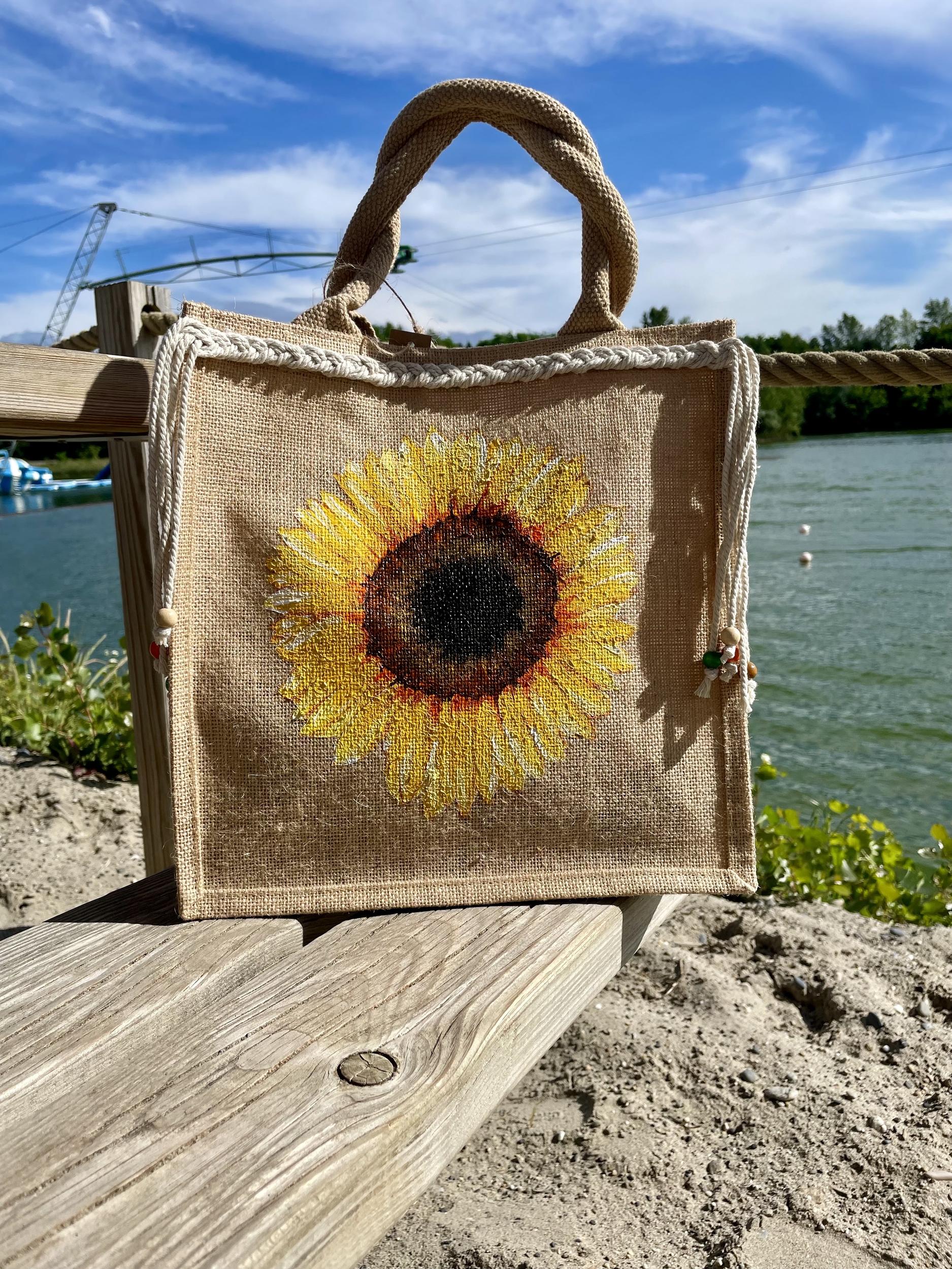 sac en jute avec un gros tournesol peint à la main par Elles et No. et décoré avec une tresse en macramé, présenté au bord de l'eau