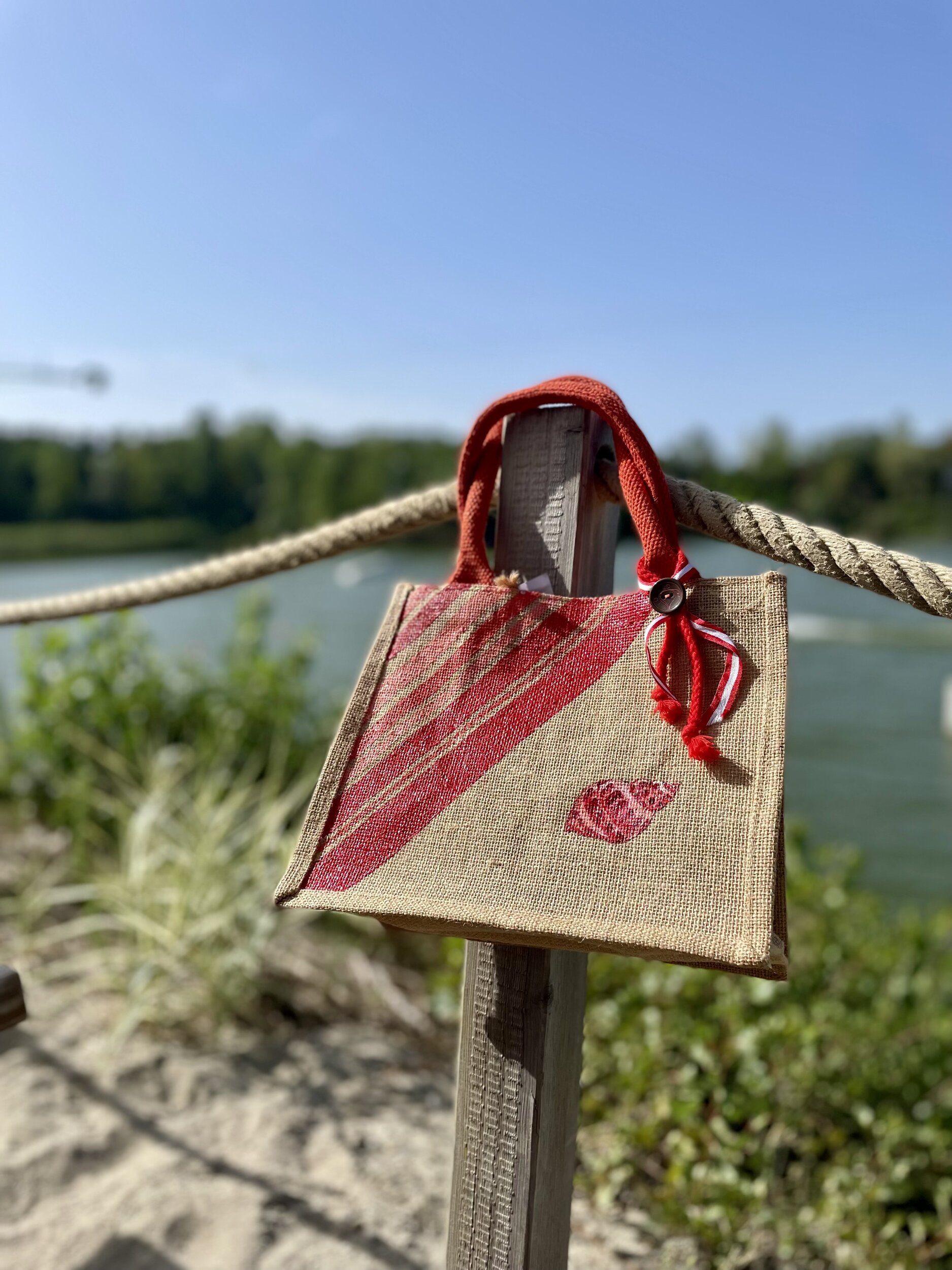 sac de plage en jute avec des rayures bayadère dans les tons rouges et un coquillage, peints à la main par Elles et No., présenté au bord de leau