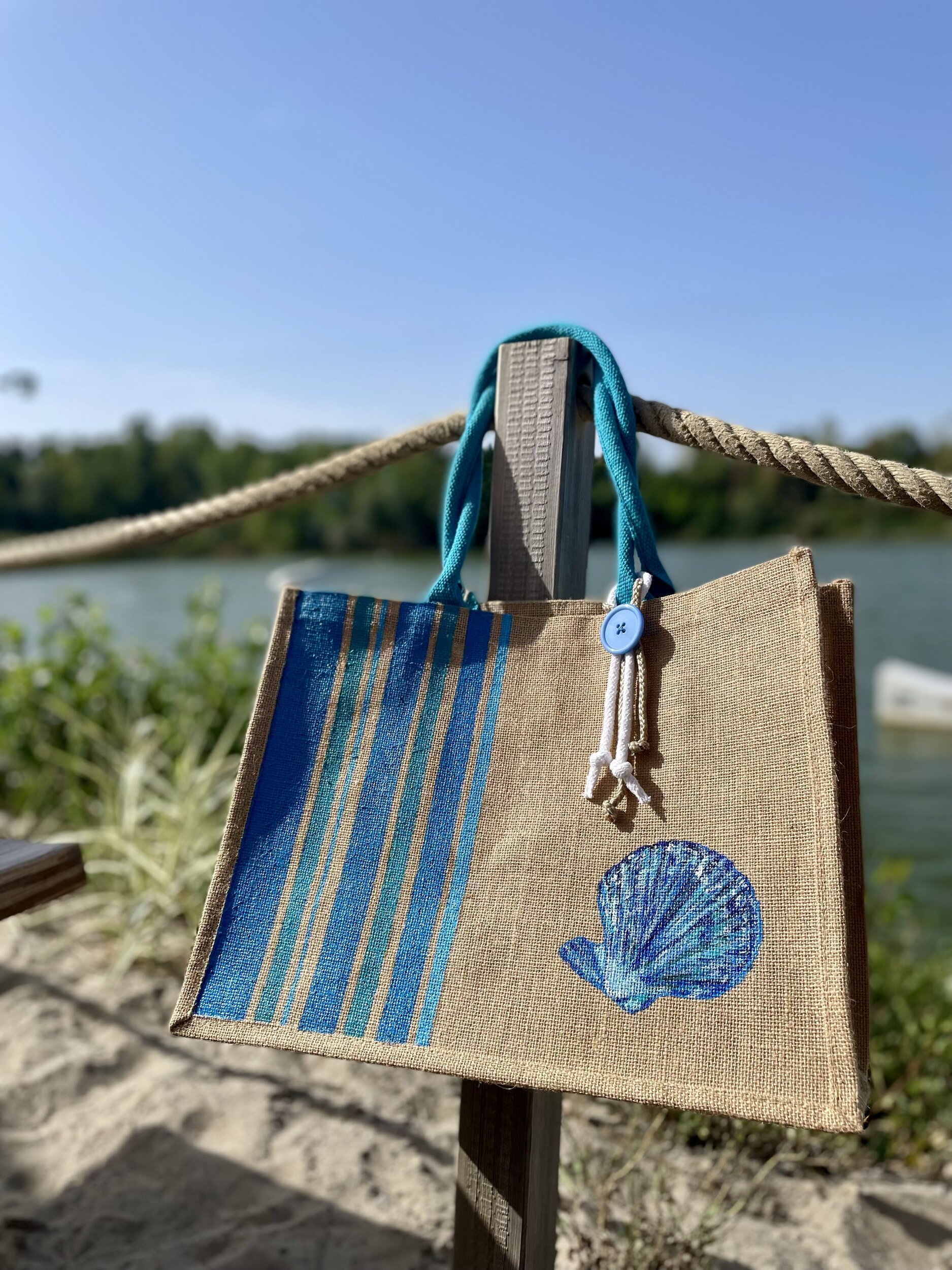 sac de plage en jute avec des rayures bayadère dans les tons bleus et un coquillage peints à la main par Elles et No., présenté au bord de leau