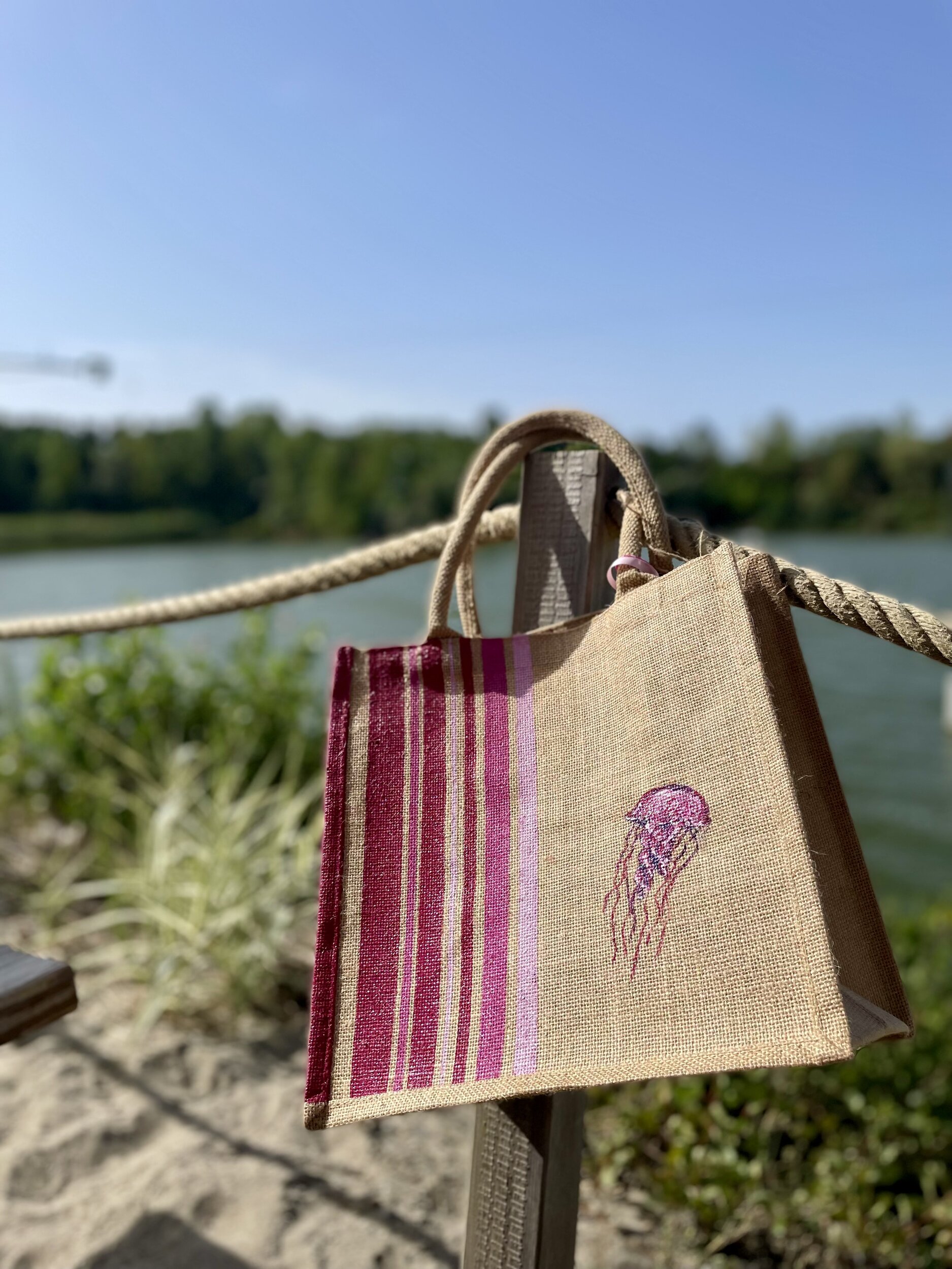 sac de plage en jute avec des rayures bayadère dans les tons roses et une méduse, peints à la main par Elles et No., présenté au bord de leau