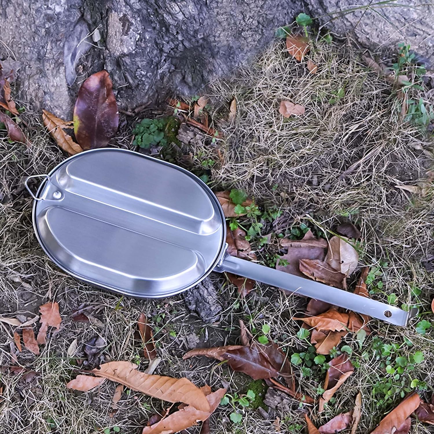 Gamelle militaire en inox posée sur un sol de forêt, manche déplié