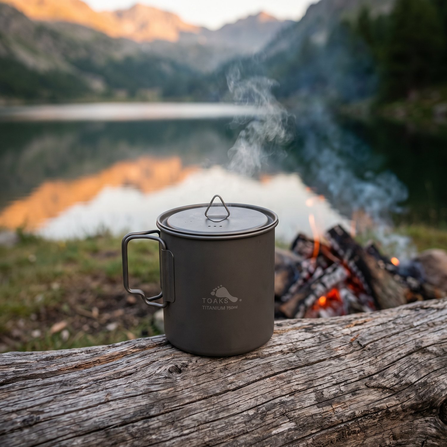 Pot en titane TOAKS 750 ml avec couvercle sur un rondin près du feu, lac de montagne au lever du jour