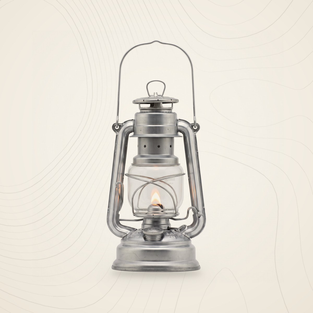 Lampe tempête Feuerhand Baby Special 276 en acier galvanisé, verre transparent et anse, sur fond lin