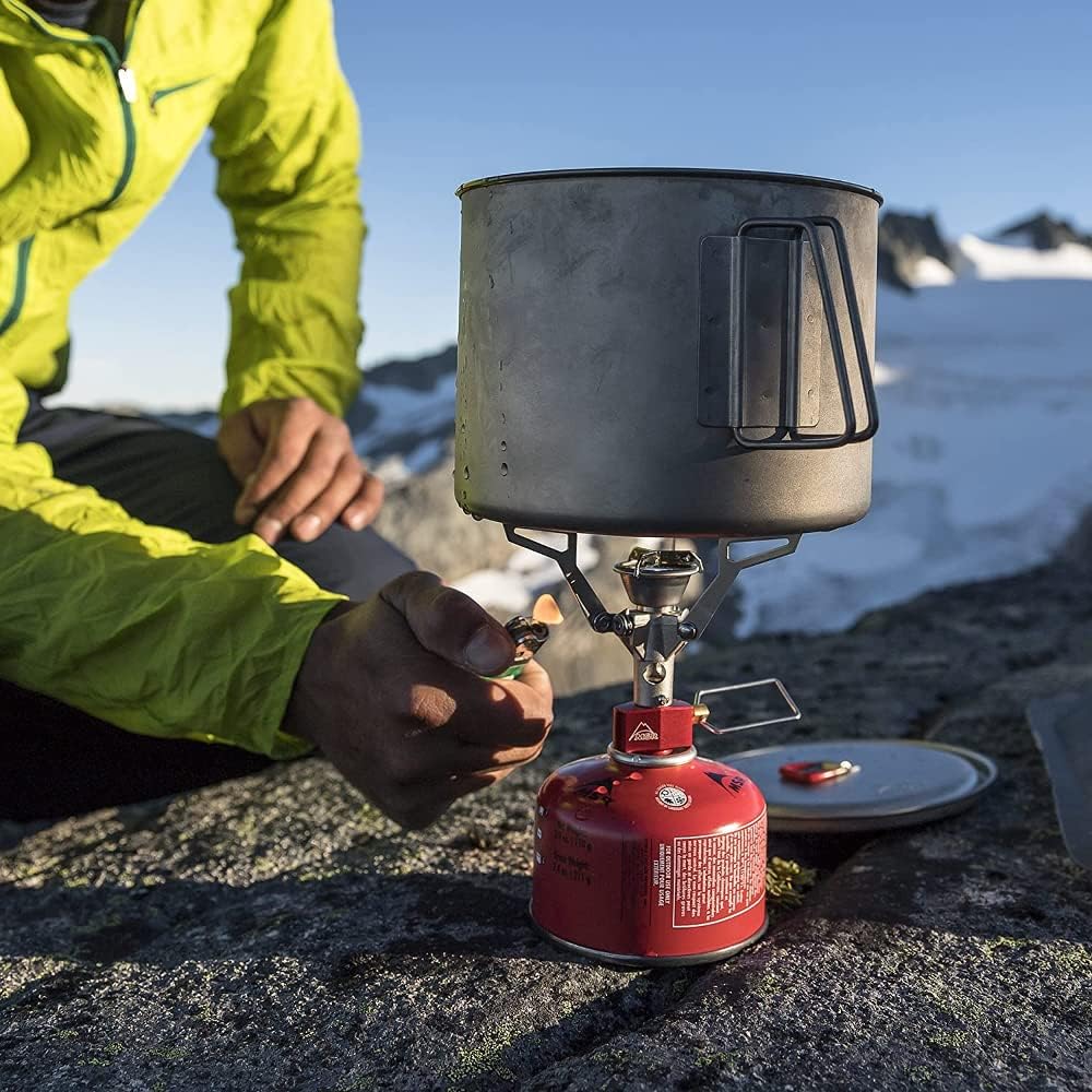 Randonneur accroupi près du PocketRocket 2 et de sa popote, sur un rocher face à un glacier
