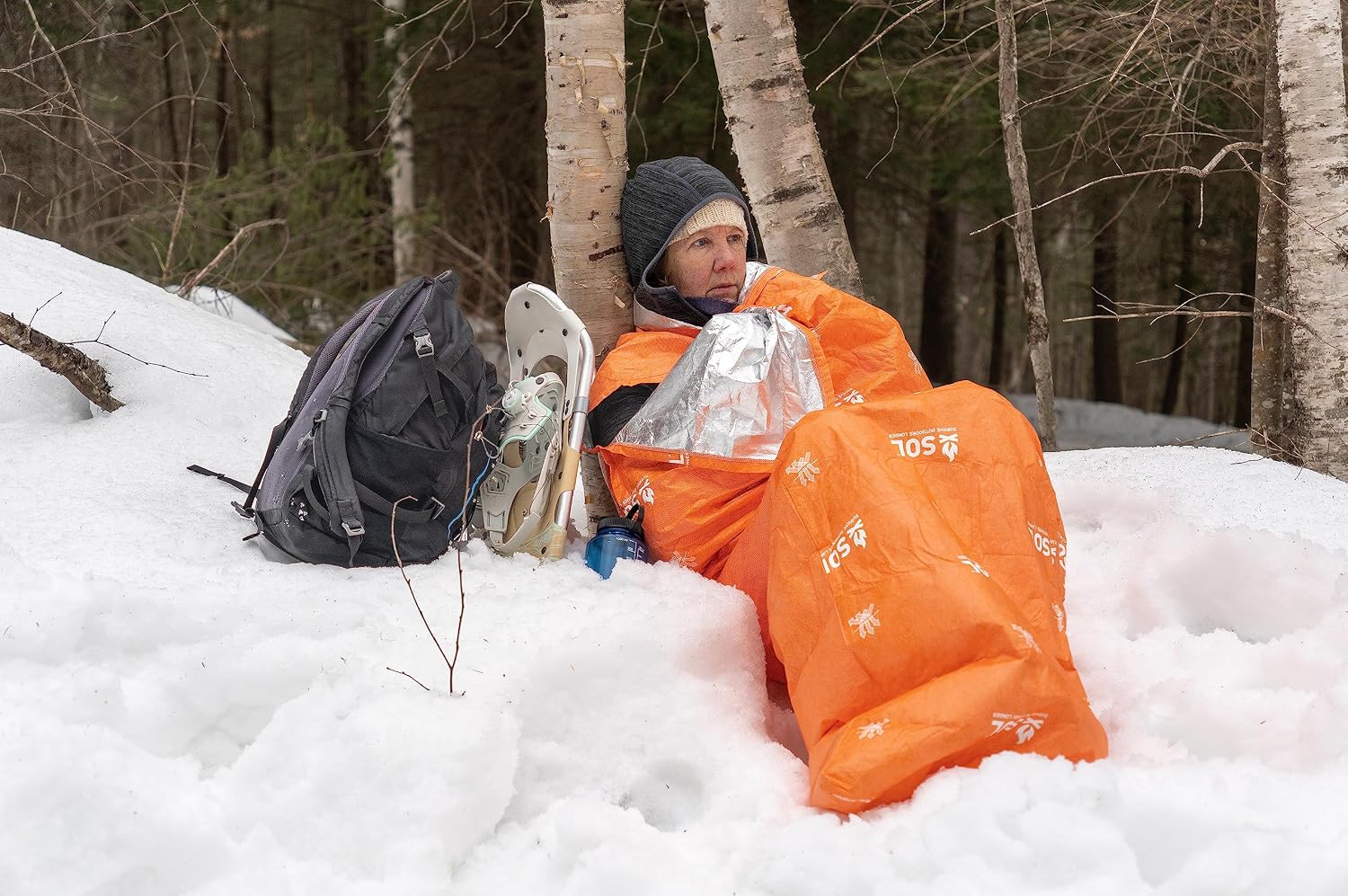 Randonneuse assise contre un bouleau, enveloppée dans le sac de bivouac SOL Escape Lite orange