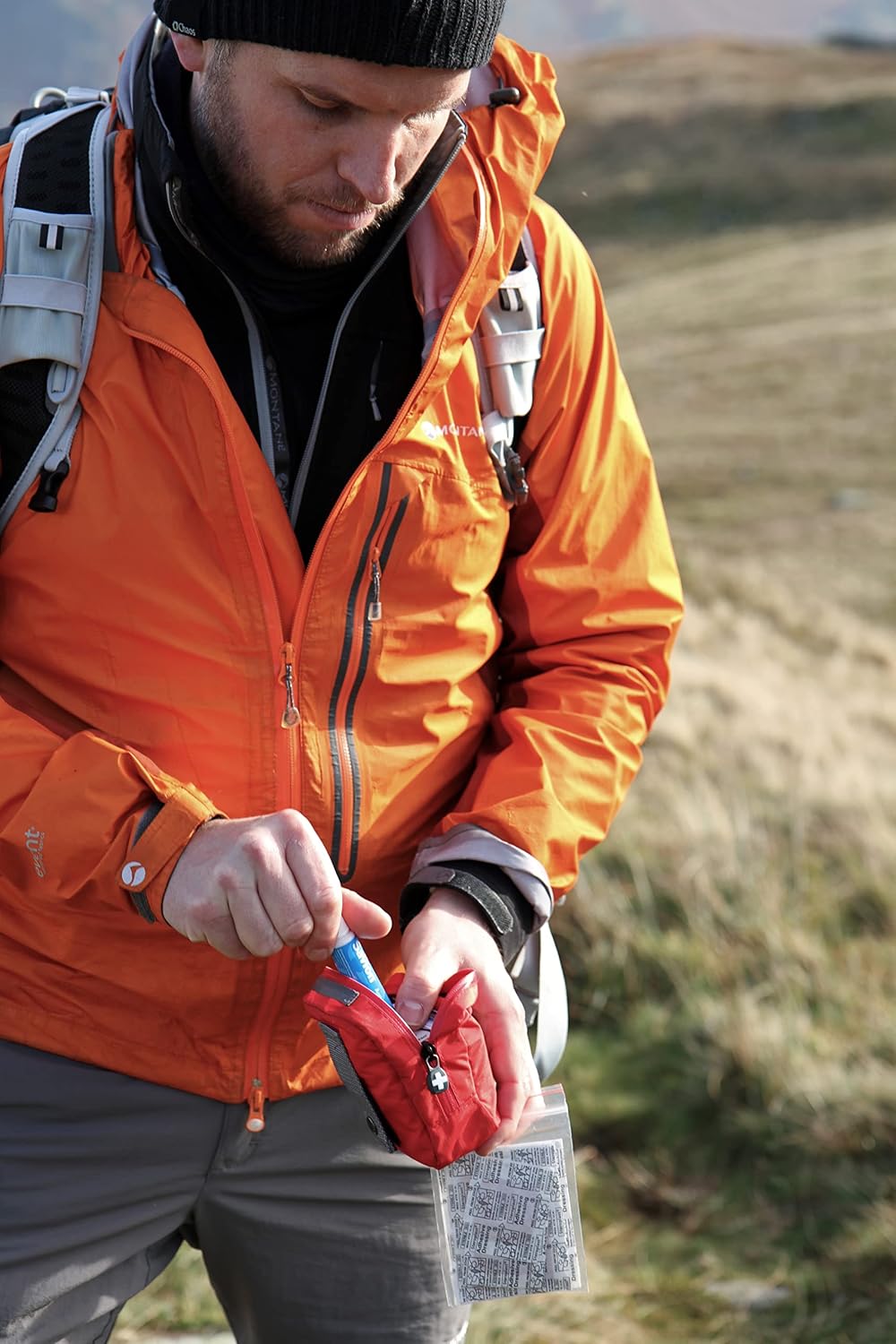 Randonneur en veste orange ouvrant la trousse Lifesystems Pocket sur une lande herbeuse