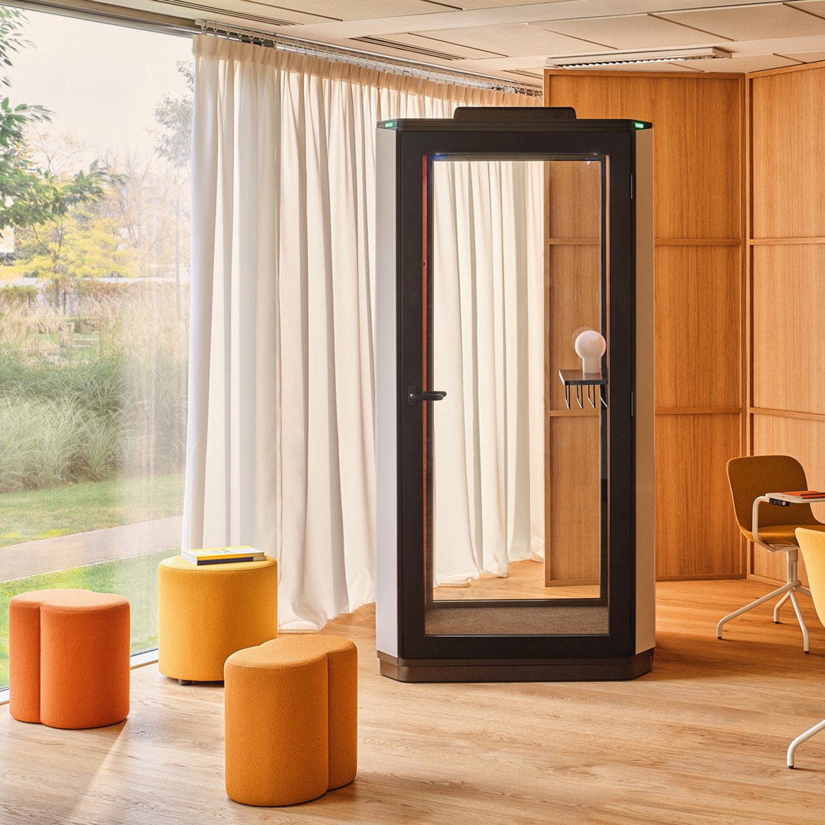 Cabine téléphonique vitrée beige et noire devant une baie vitrée, poufs orange au premier plan