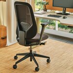 Chaise de bureau ergonomique noire à dossier résille devant un bureau réglable, près d'une fenêtre