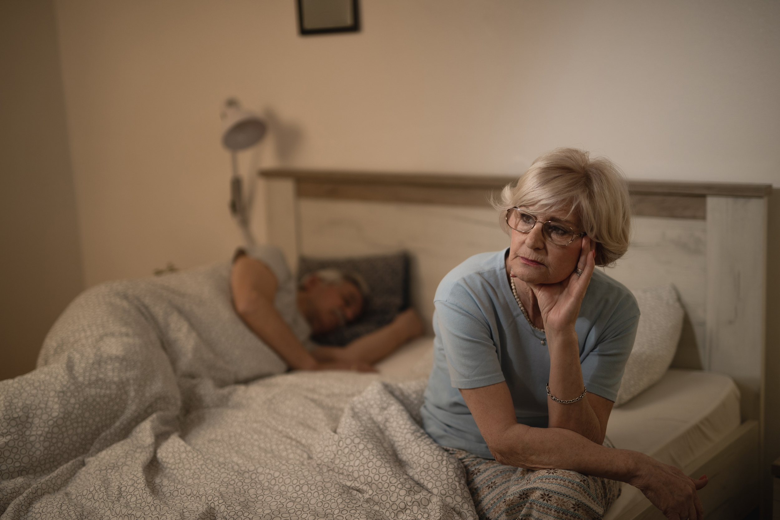 pensive mature woman feeling sleepless and sitting in the bed while her husband is sleeping in the background