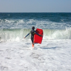 Jeune adolescent de dos en combinaison et planche de surf rouge qui marche vers les vagues