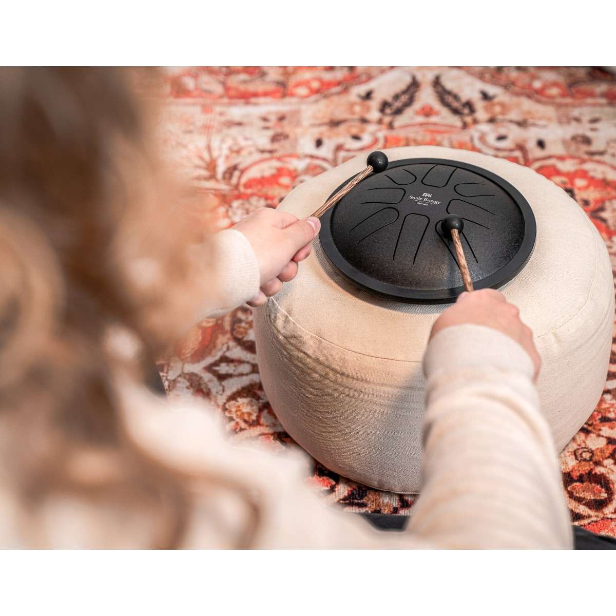 Personne jouant du mini steel tongue drum Meinl noir avec des maillets, posé sur un coussin rond beige