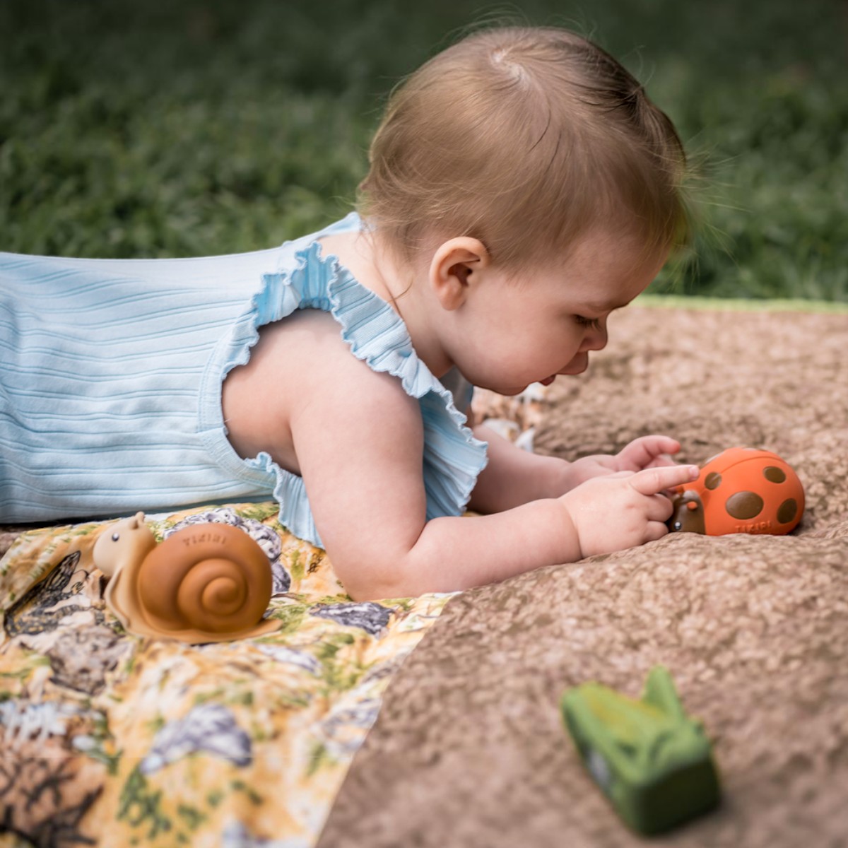 Bébé à plat ventre touchant la coccinelle Tikiri, escargot en caoutchouc naturel posé à côté delle