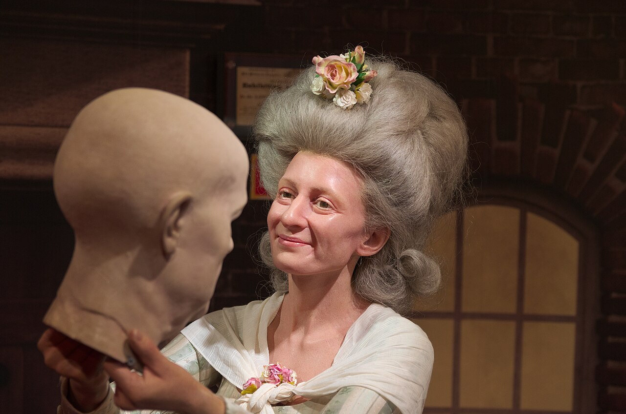 Statue de cire de Marie Tussaud sculptant un buste, coiffure 18ème siècle et fleurs, sur la page Chroniques de Cire