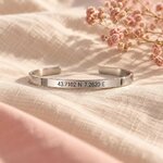 Bracelet jonc argenté gravé de coordonnées GPS posé sur un lin crème