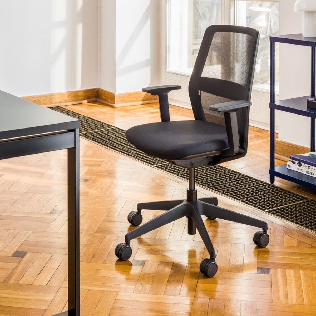 Chaise de bureau à dossier résille noire avec accoudoirs et piétement noir sur un parquet