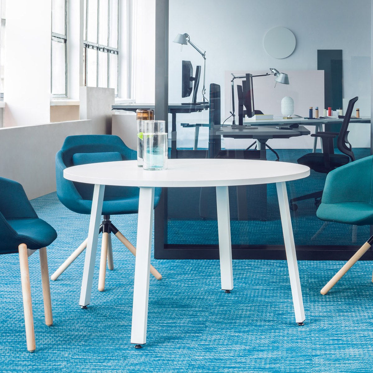 Table ronde Ogi A blanche à pieds blancs inclinés entre des fauteuils bleus dans un open space