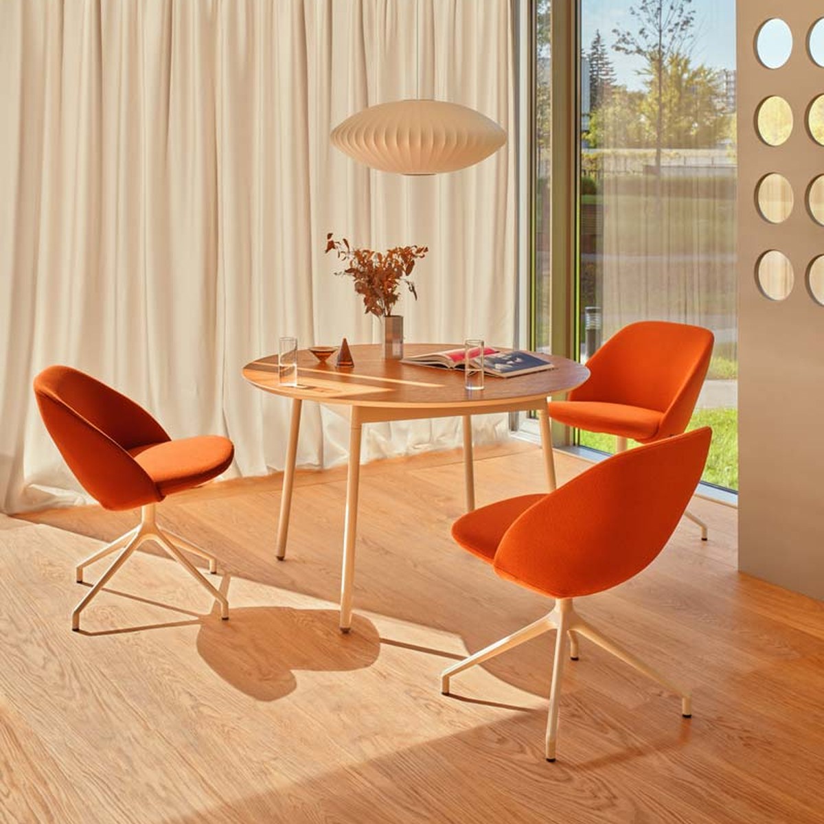 Table ronde Unit en chêne cuivré à pieds blanc perle avec trois fauteuils orange près dune baie vitrée