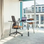 Chaise de bureau à dossier résille noir et assise orangée devant une table, près dune baie vitrée