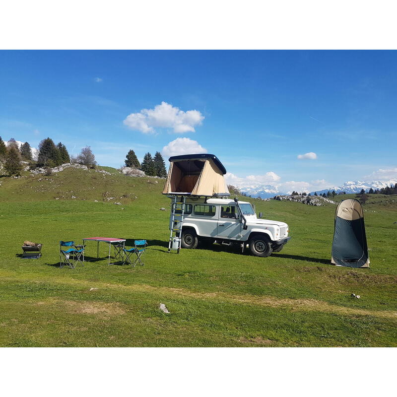 Tente de toit NaïtUp ouverte sur un 4x4 blanc dans une prairie de montagne, table et chaises