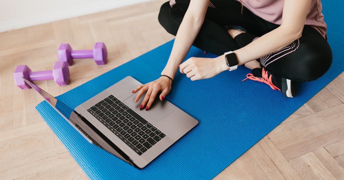 crop female in sportswear sitting on blue yoga mat on floor and surfing internet on laptop