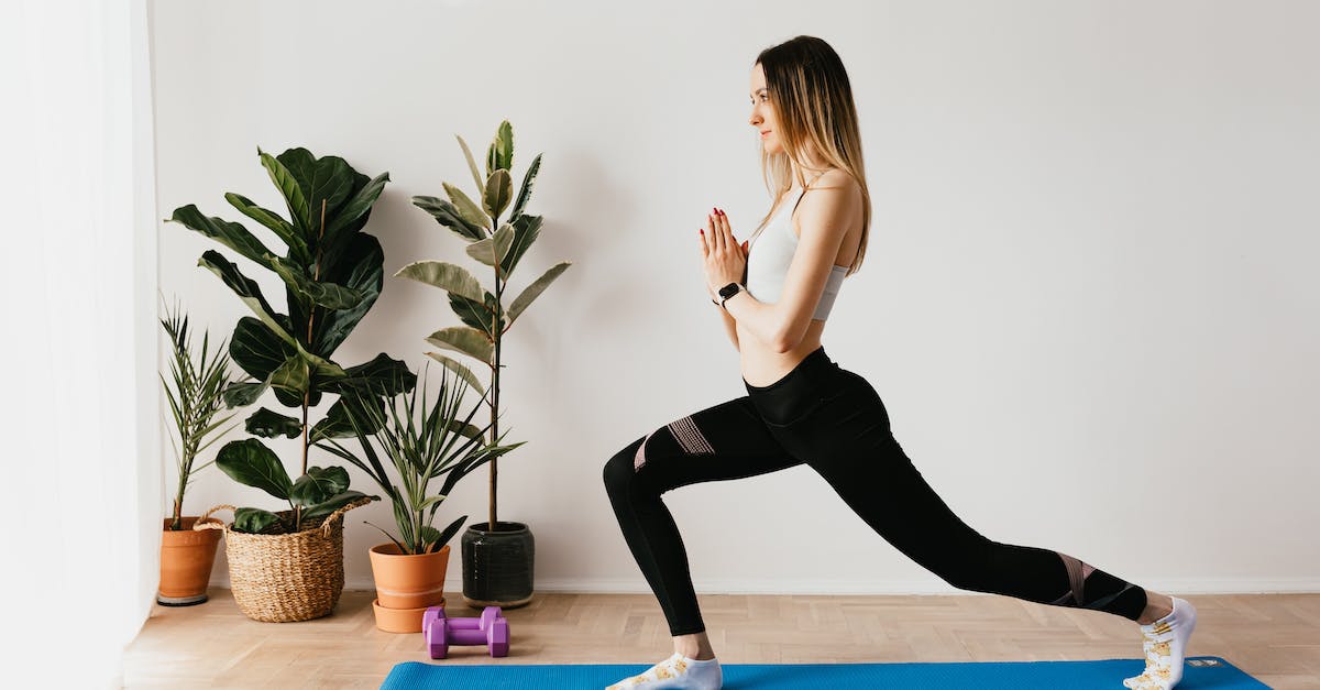side view of fit lady in sports clothes and wristwatch standing in anjaneyasana pose while practicing yoga on mat near dumbbells and potted plants at home and looking away