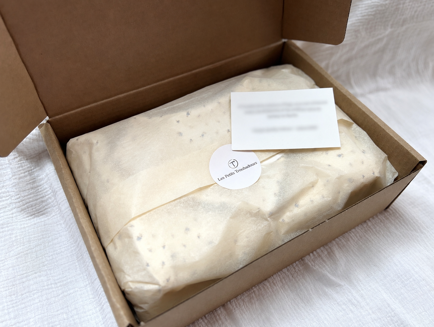 Coffret de naissance emballé dans du papier de soie, avec une carte et l'autocollant Les Petits Troubadours