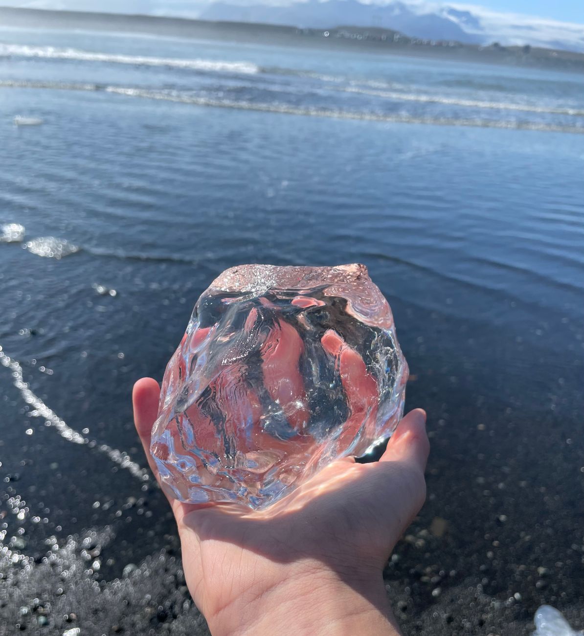 un morceau d iceberg transparent maintenu sur du sable noir avec l ocean en arriere plan sous un ciel bleu clair