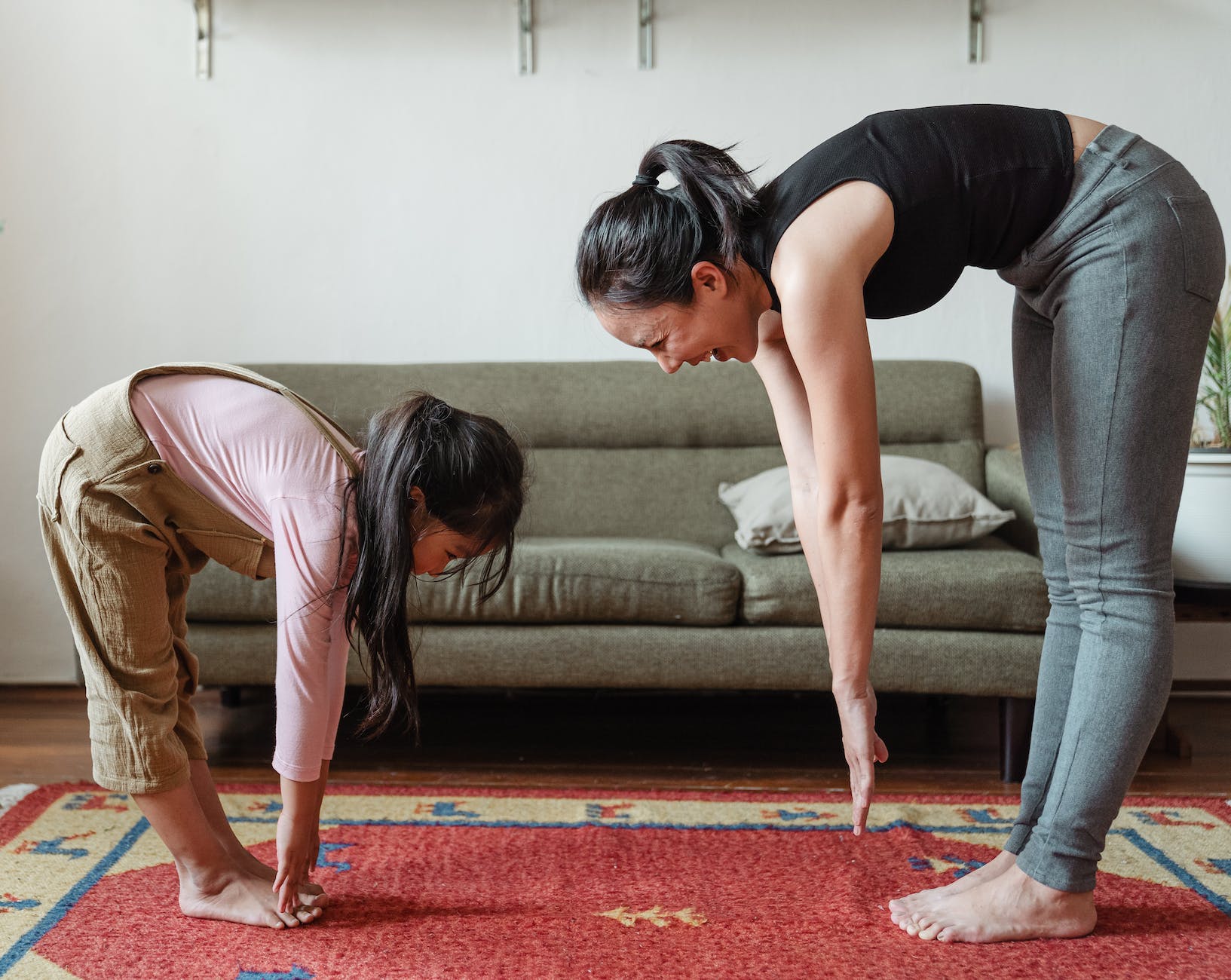 side view of smiling young asian woman stretching body with adorable daughter while standing against each other on floor carpet near cozy couch in modern living room in morning
