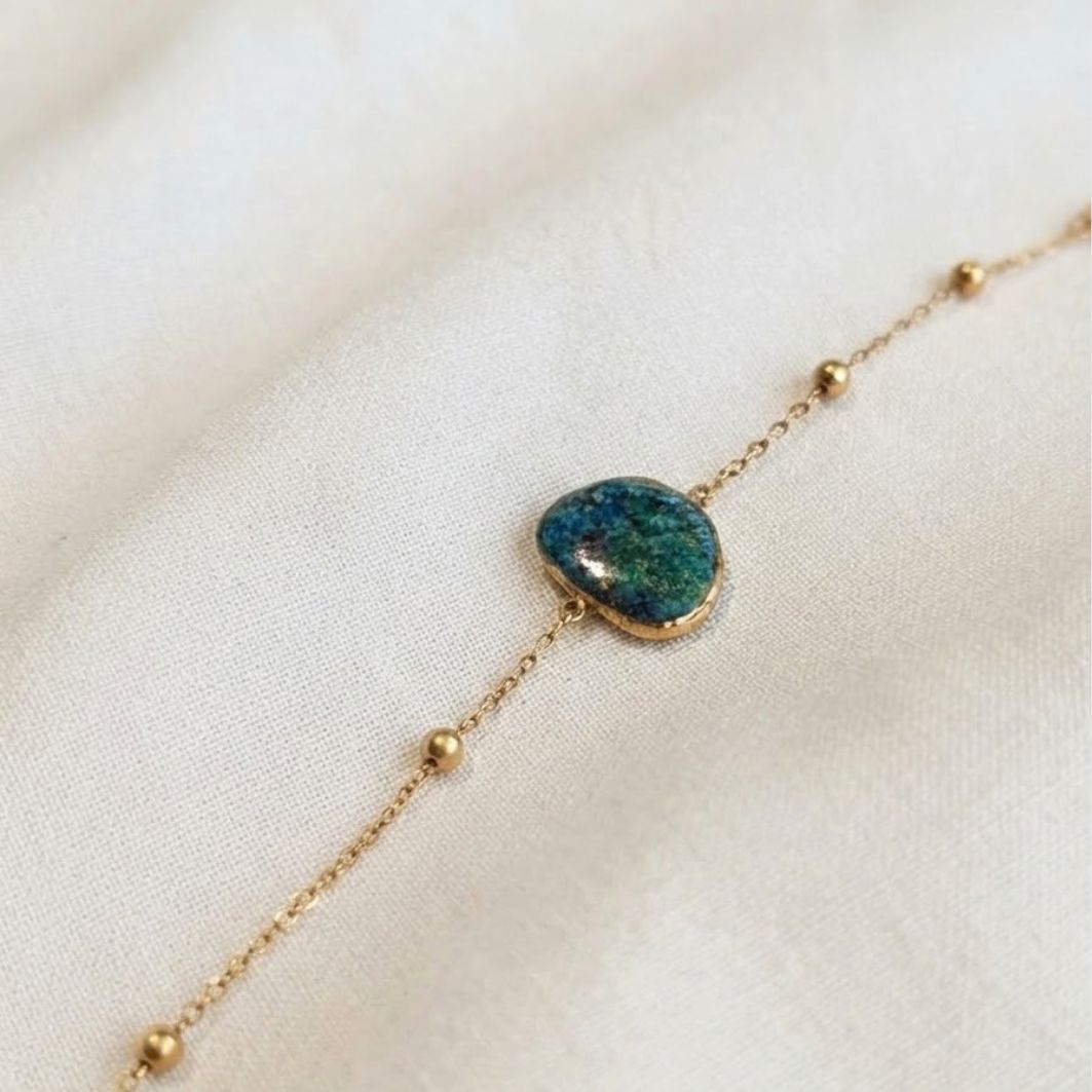 Bracelet Nour, chaîne à billes dorées et pierre en béton irisé bleu-vert, posé sur lin