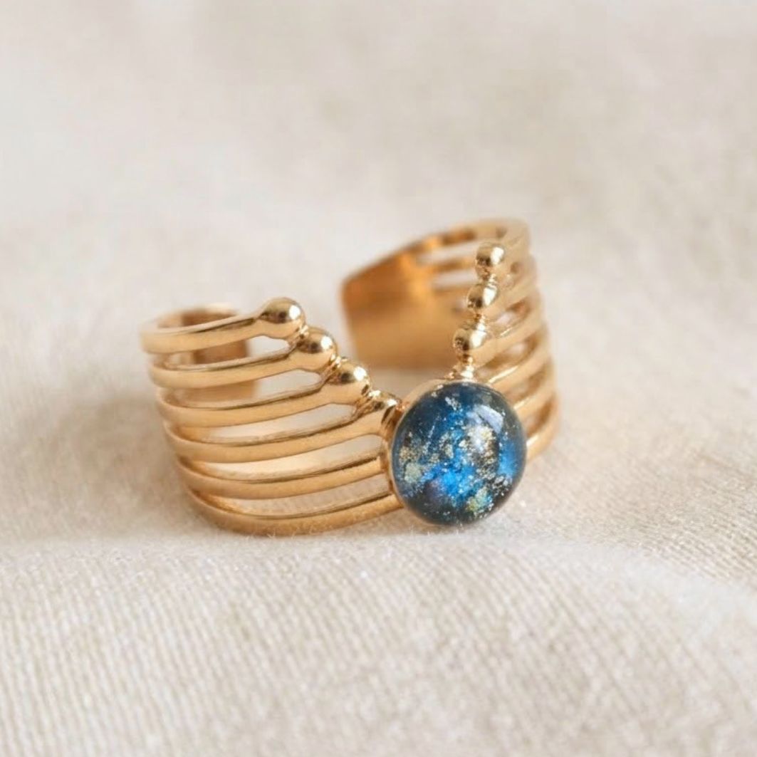 Bague Cléopâtre en acier inoxydable doré, bandes ajourées à billes et pierre en béton irisé bleu-vert, posée sur lin