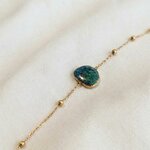 Bracelet Nour, chaîne à billes dorées et pierre en béton irisé bleu-vert, posé sur lin