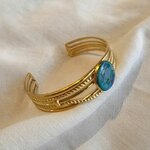 Jonc Méret ajustable en acier inoxydable doré, détails cordés et pierre en béton irisé bleu-vert, posé sur lin